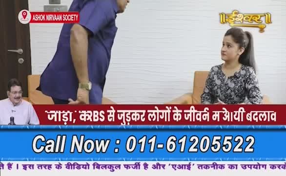 A man in a blue shirt is moving to the left, while a woman on Ishwar Bhakti TV looks toward him. The lower third of the screen displays a phone number and text in Hindi.
