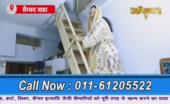 A woman in a patterned robe is climbing a wooden ladder, her head covered. The scene appears to be from a broadcast on Ishwar Bhakti TV in India, with a phone number displayed at the bottom.
