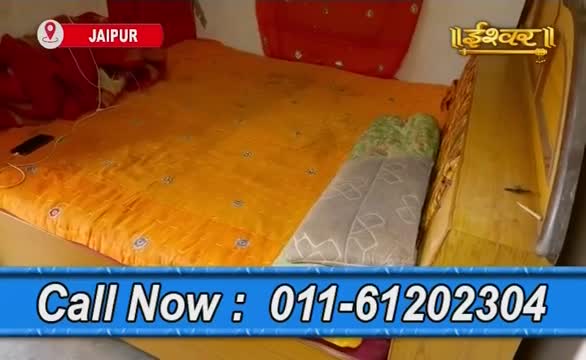 A bed covered in a bright yellow cloth fills the frame, with a patterned pillow and a red cushion visible. The Ishwar Bhakti TV logo sits in the top right corner, and a phone number flashes at the bottom.
