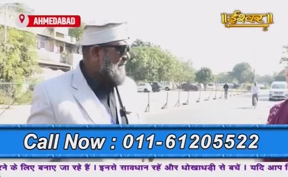 A man in a white suit, wearing sunglasses and a white cap, is speaking on a street in Ahmedabad, India. The Ishwar Bhakti TV logo is in the corner, and a phone number is displayed on the screen.
