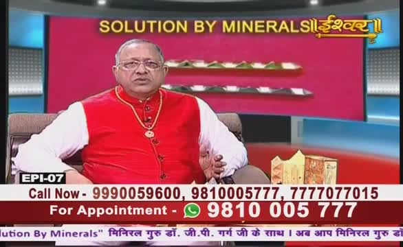 The man in the red vest and glasses gestures while speaking, likely on Ishwar Bhakti TV in India. Below, phone numbers are displayed for appointments with "Solution by Minerals."
