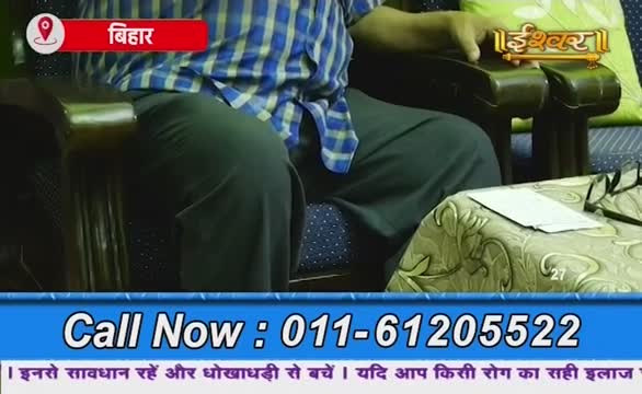 A man in a blue and white checked shirt sits in a chair, the Ishwar Bhakti TV logo visible in the corner. Below, a phone number flashes on screen, with a message about avoiding fraud in Hindi.
