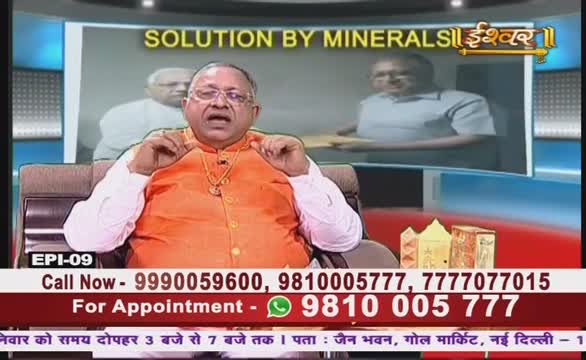A man in an orange tunic gestures with his hands while speaking on a television program from India, likely on Ishwar Bhakti TV. Below him, phone numbers and appointment information are displayed on the screen.
