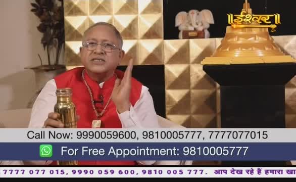 The man in the red vest gestures with his right hand while holding a small, gold-colored bottle. He's on what looks like an Ishwar Bhakti TV program in India, with a golden structure and a small elephant statue in the background.
