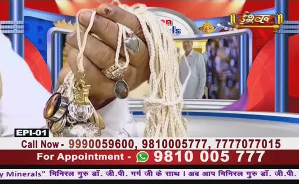 A hand, adorned with rings, displays a collection of pendants and beads on the Ishwar Bhakti TV set. A man in a light-colored shirt is visible in the background.
