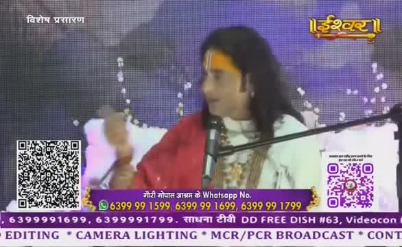 A man, wearing a red shawl, speaks into a microphone on a set for Ishwar Bhakti TV. The screen behind him displays the channel's logo and contact information.
