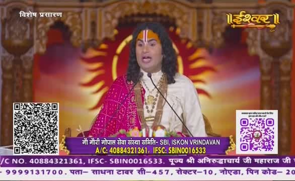 A man in a white and pink outfit speaks into a microphone, his face animated as he addresses the audience. The scene is broadcast on Ishwar Bhakti TV, with the logo visible in the upper right corner.
