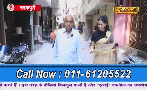 A man and a woman walk down a narrow street in India, with a motorcycle parked on the side. The woman, holding a container, is likely part of a segment on Ishwar Bhakti TV.
