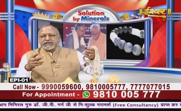 A man in a tan vest gestures with one hand while holding a string of beads in the other. The Ishwar Bhakti TV screen behind him shows a bracelet, and a banner advertises free consultations in Hindi.
