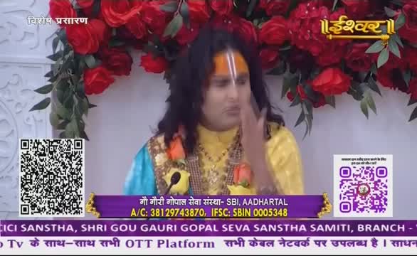 A man in a yellow and blue garment gestures with his right hand, a white mark on his forehead. The scene is from India, likely a broadcast on Ishwar Bhakti TV, with floral decorations and a QR code visible.
