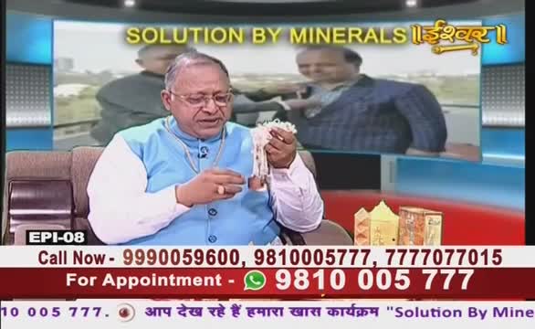 A man in a blue vest holds a bundle of white objects, likely for display on Ishwar Bhakti TV. Behind him, a screen shows a blurry image, and a phone number scrolls across the bottom for appointments.
