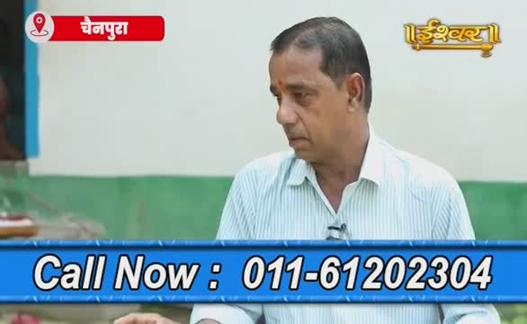 The man in the light blue shirt is looking down, seemingly listening intently. A phone number flashes across the bottom of the screen, suggesting a call-in segment on Ishwar Bhakti TV.
