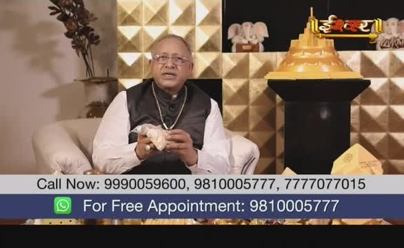 A man in a black vest and glasses sits on a cream-colored couch, holding a small, off-white object. The Ishwar Bhakti TV logo is visible behind him, and phone numbers scroll across the bottom of the screen.
