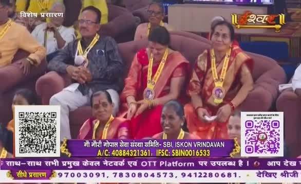 People sit on a plush, curved couch, adorned with yellow sashes and medals, likely part of a broadcast on Ishwar Bhakti TV in India. The image is framed by text and QR codes, advertising the event.

