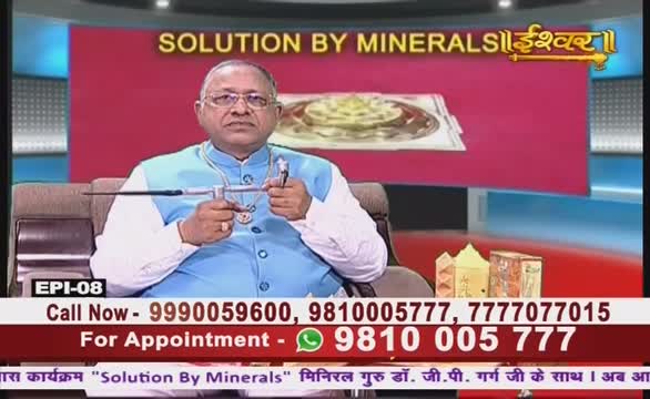 A man in a blue vest sits on a chair, holding a small, metal device. He's on a TV set, with a red background and the Ishwar Bhakti TV logo visible above him.

