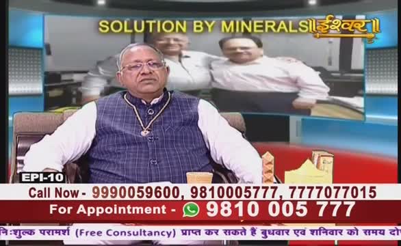 A man in a blue vest, wearing glasses and a pendant, sits in a chair on a set for Ishwar Bhakti TV. The screen behind him displays a graphic about minerals, and a phone number scrolls at the bottom.
