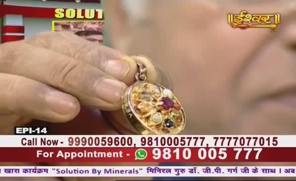 A hand holds up a gold pendant, studded with colorful stones, on what looks like an Ishwar Bhakti TV show. The bottom of the screen displays phone numbers for appointments, with a "Solution By Minerals" program title.
