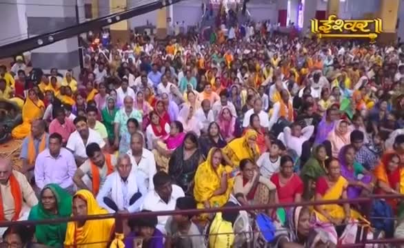 A vast crowd sits on the floor, a sea of colorful clothing filling the space. The Ishwar Bhakti TV logo sits above the gathering, likely filming an event in India.
