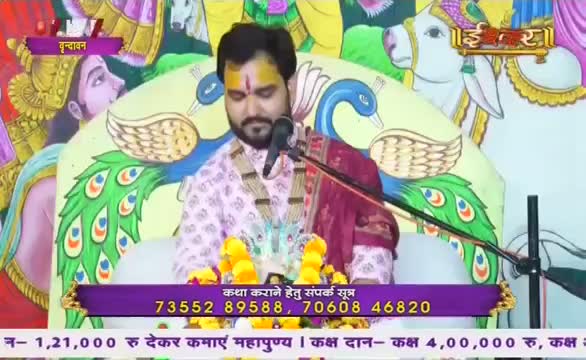 A man in a patterned shirt and maroon shawl sits on a white cushion, speaking into a microphone. The scene is from Ishwar Bhakti TV, with peacock and cow imagery in the background.

