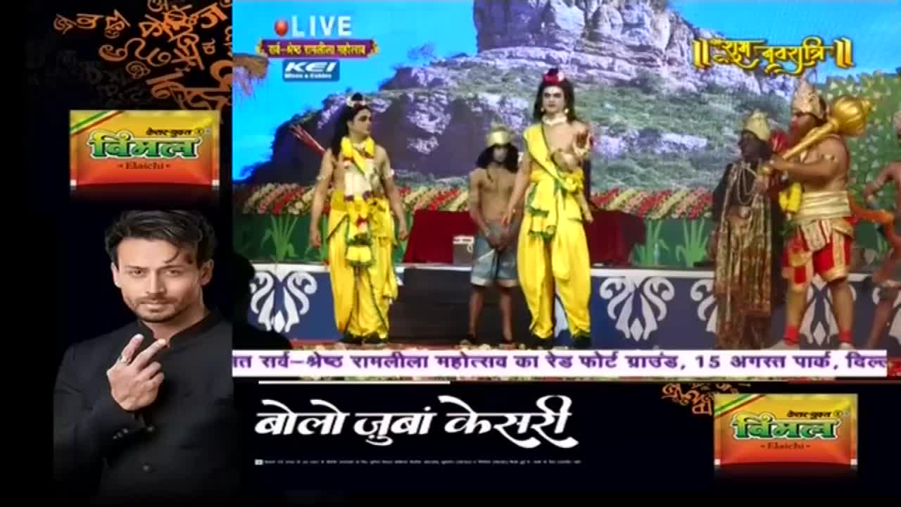 On stage, performers in vibrant yellow costumes portray characters from a religious drama, broadcast live on Ishwar Bhakti TV. A man in a suit gestures toward the scene, while advertisements for Vimal products frame the action.
