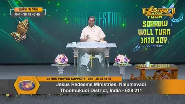 A man in a light-colored shirt stands behind a podium, gesturing as he speaks. The backdrop of the Ishwar Bhakti TV program includes the words "Sorrow Will Turn Into Joy" and a phone number for prayer support from India.
A man in a light-colored shirt stands behind a podium, gesturing as he speaks. The backdrop of the Ishwar Bhakti TV program includes the words "Sorrow Will Turn Into Joy" and a phone number for prayer support from India.
