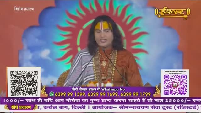 A man in traditional Indian attire speaks into two microphones, his face lit by the studio lights. The broadcast, likely from India's Ishwar Bhakti TV, features a backdrop of a sun-like design.
