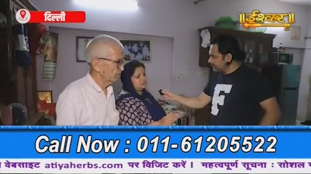 A man in a black t-shirt holds a microphone, speaking to an older couple in their home. The Ishwar Bhakti TV logo is visible in the corner, and a phone number flashes at the bottom of the screen.
