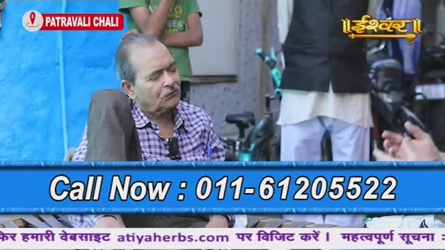 A man in a checkered shirt sits with his legs crossed, looking to his right. The Ishwar Bhakti TV logo and a phone number are displayed at the bottom of the screen.
