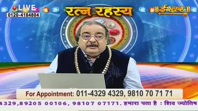 A man in a navy vest and pearl necklace sits at a desk, speaking on Ishwar Bhakti TV. The screen behind him displays the show's title, "Ratan Rahasya," and a phone number for appointments.
