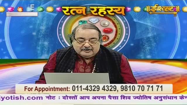A man in glasses sits behind a desk, facing the camera on Ishwar Bhakti TV. He's wearing a dark vest over a red shirt, and a laptop sits in front of him.
