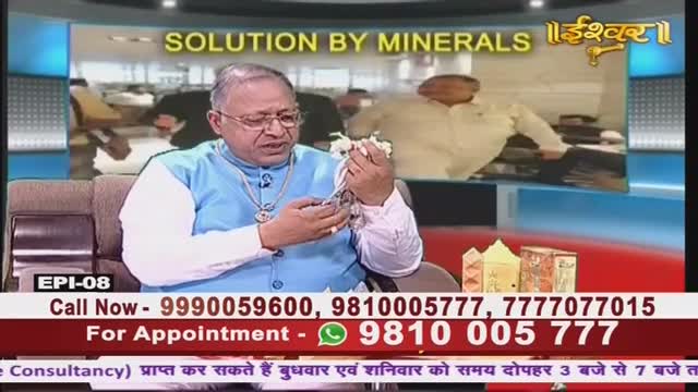 The man on the Ishwar Bhakti TV program in India holds a small, white object in his hands, gesturing as he speaks. Behind him, a screen displays the words "Solution by Minerals" and a blurry image of other people.
