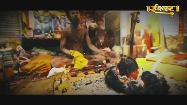 A man in yellow cloth sits amidst offerings, tending to a small fire. Flames lick upwards from a dark, charred object in the foreground.