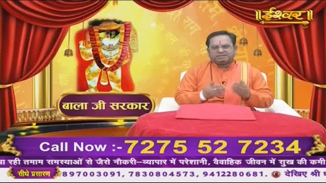 A man in an orange robe speaks with his hands gesturing, seated before a large red cushion. Behind him, a colorful image of a deity is displayed, framed by red curtains.