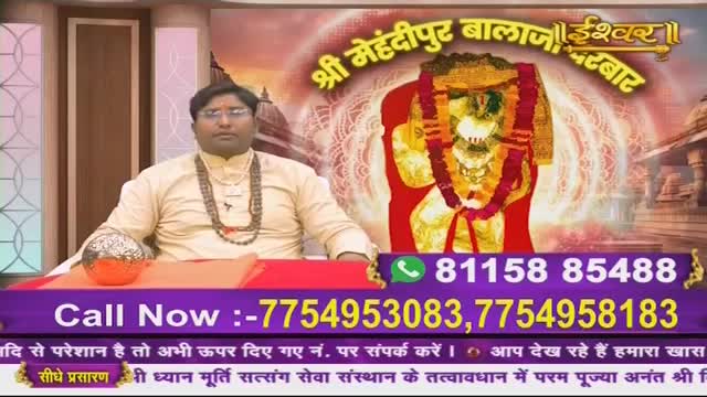 A man in a light yellow kurta sits at a table, a beaded necklace around his neck. Behind him, a vibrant image of a deity is displayed on a screen, with text in Hindi and phone numbers.