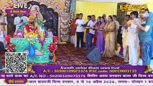 A man in a vibrant yellow turban and ornate robes sits on a decorated throne, a garland of marigolds around his neck. A crowd of people, dressed in traditional Indian attire, stand before him, some holding plates of offerings. The Ishwar Bhakti TV logo is visible in the background, indicating the live broadcast of this spiritual gathering.