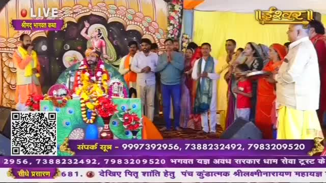 A man in a vibrant orange and yellow robe stands to the left, speaking into a microphone. In the center, a figure adorned with garlands sits on a decorated platform, surrounded by flowers and peacock imagery.