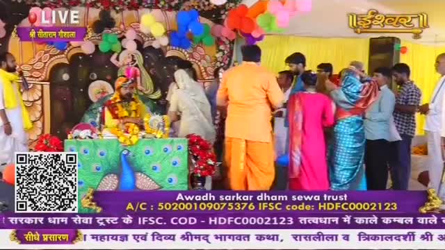 A man in a yellow dhoti and turban sits on a decorated platform, while others in vibrant saris and kurtas gather around. A large peacock design adorns the front of the stage, with red flowers adding a splash of color.