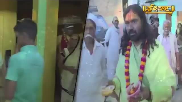 A man in a light green robe, adorned with a garland of marigolds and roses, walks forward. Behind him, others in white attire follow, some with their faces partially obscured.