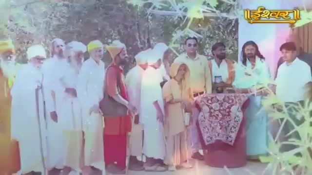 A group of men, some in white robes, stand together. A woman in a light-colored outfit stands near a decorated table. The Ishwar Bhakti TV logo is visible in the background.