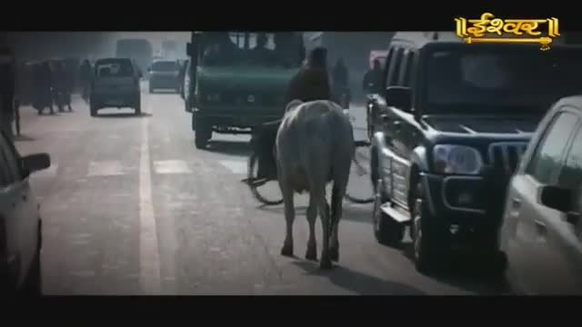 A white cow ambles down the middle of a busy Indian road, weaving between cars and a green truck. The Ishwar Bhakti TV logo is visible in the upper right corner.
