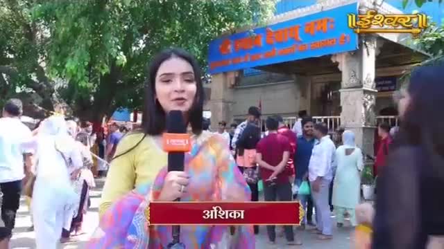 A reporter stands with a microphone outside a temple, her colorful sari draped over her arm. A crowd of people, dressed in a mix of traditional and modern Indian attire, mills about behind her.