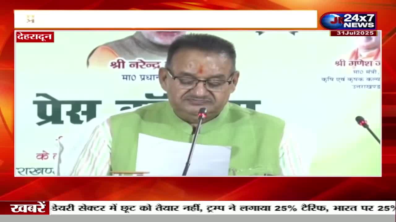 A man in glasses and a green vest speaks into a microphone, reading from a paper. The JK 24x7 News logo is visible, and the backdrop indicates a press conference in Dehradun, India.
