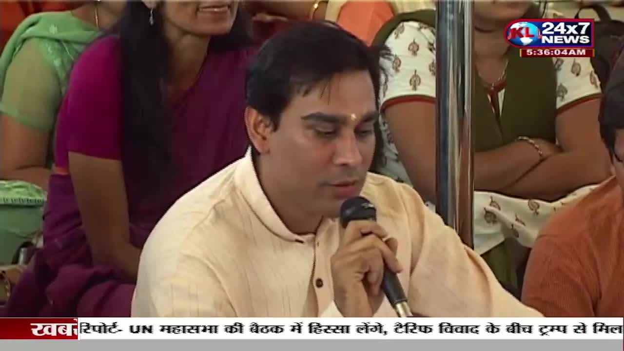A man in a light-colored shirt speaks into a microphone, likely on a news program from India, as the JK 24x7 News logo is visible in the corner. The background is filled with people, some wearing traditional Indian clothing.
