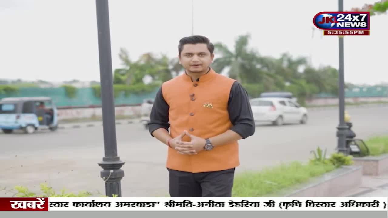 A man in an orange vest stands in front of a camera, speaking. Behind him, cars and a blue auto-rickshaw move on the road, as seen on JK 24x7 News.
