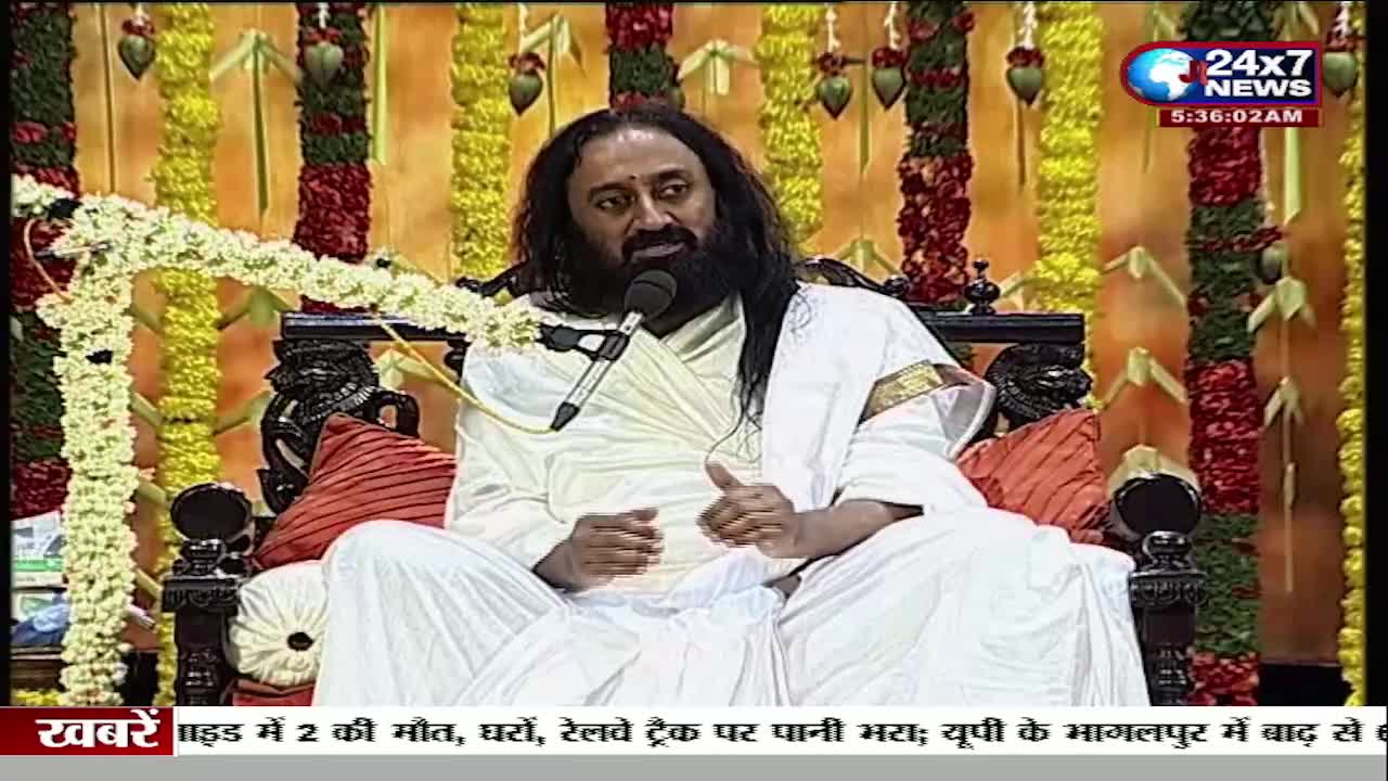 A man in white robes sits on a decorated chair, speaking into a microphone. The JK 24x7 News logo is visible, and the news ticker at the bottom mentions flooding and deaths in India.
