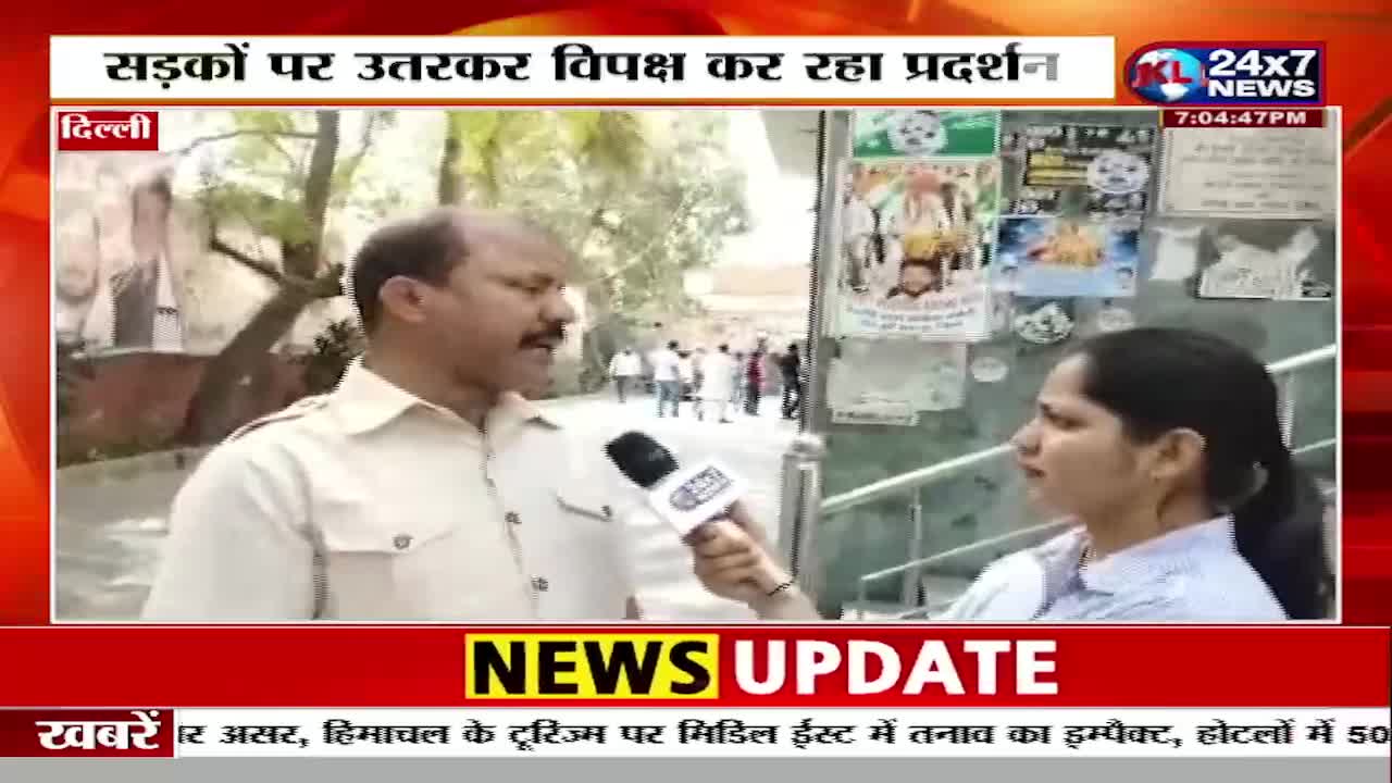 A man in a light-colored uniform speaks into a microphone held by a woman. Behind them, a group of people are visible in the distance, and posters are plastered on a wall.