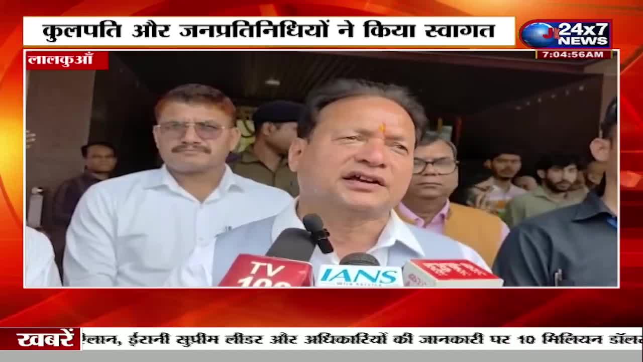 A man in a light blue shirt speaks into a microphone, flanked by other men. Behind them, a news graphic from 24x7 News displays text in Hindi.
