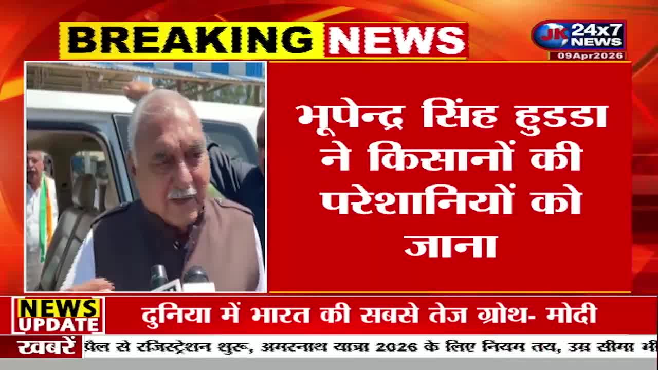 A man in a brown vest and white shirt speaks into microphones, his white hair and mustache prominent. Behind him, a white SUV is parked, and a flag with green, white, and saffron stripes is visible inside. A man in a brown vest and white shirt speaks into microphones, his white hair and mustache prominent. Behind him, a white SUV is parked, and a flag with green, white, and saffron stripes is visible inside.