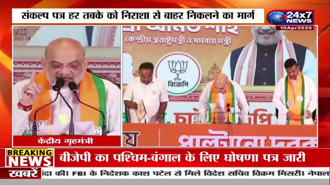 The Union Home Minister speaks at a podium, his hand raised in emphasis. Beside him, other men in white shirts and orange sashes stand behind a table, one holding a document. A large portrait of a man in a suit and saffron scarf looms behind them. The Union Home Minister speaks at a podium, his hand raised in emphasis. Beside him, other men in white shirts and orange sashes stand behind a table, one holding a document. A large portrait of a man in a suit and saffron scarf looms behind them.