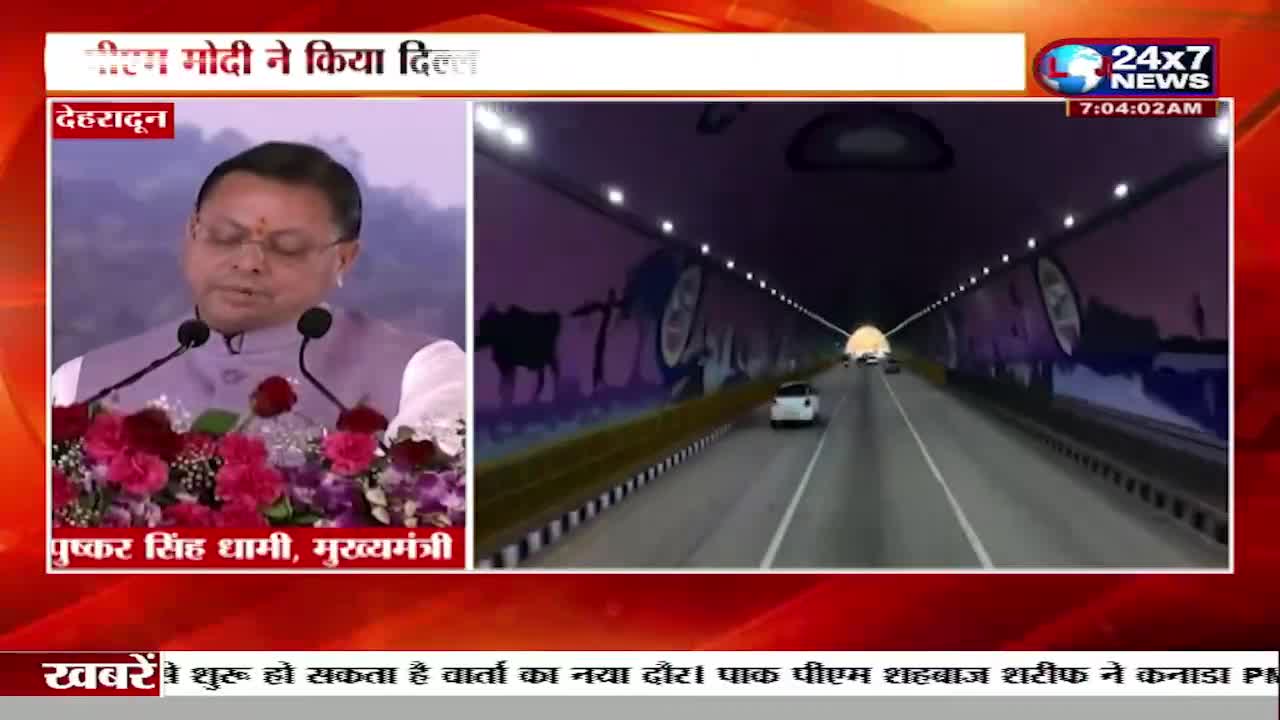 A man in a light purple jacket speaks at a podium, flanked by microphones and a vibrant floral arrangement. Ahead, cars travel through a brightly lit tunnel adorned with murals of cows and rural scenes.